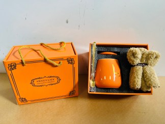 Lote #018 - Dos cajas para regalería que c/u contiene: una taza en cerámica esmaltada de color + cuchara color oro + toalla pequeña (color naranja)