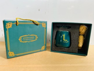 Lote #019 - Dos cajas para regalería que c/u contiene: una taza en cerámica esmaltada de color + cuchara color oro + toalla pequeña (color verde)